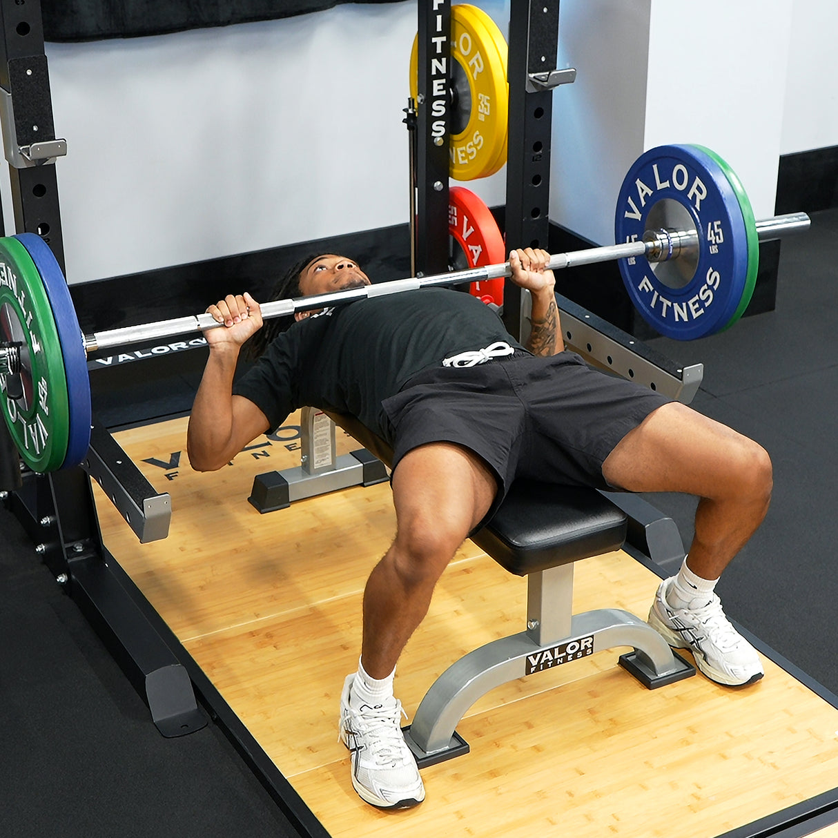 Person performing bench press with Valor Fitness flat bench, barbell, plates, and half rack in a gym setting